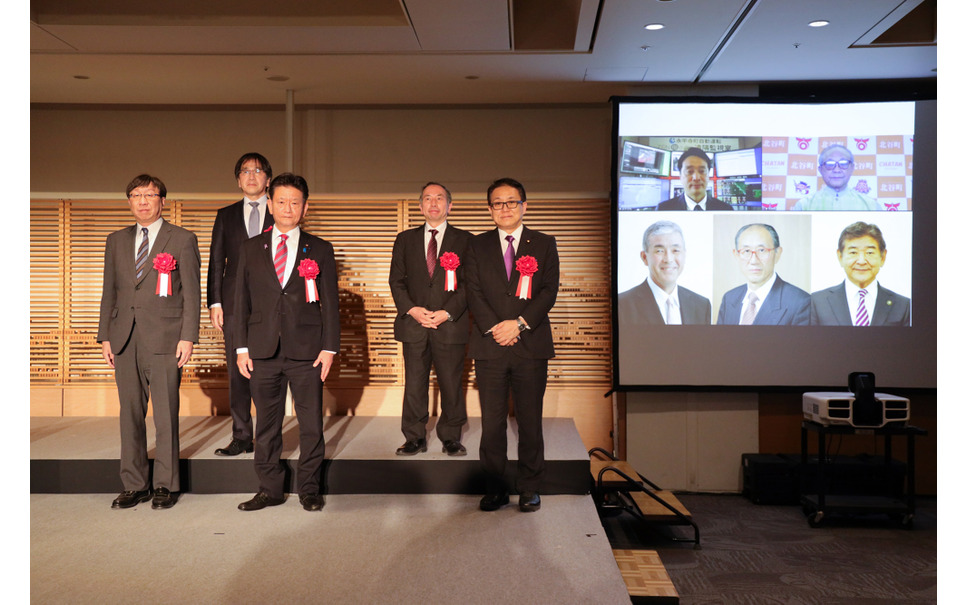 前列右より、経産省・江島潔副大臣、内閣府・三ツ林裕巳副大臣、国交省・渡辺学審議官。後列右より、東京大学 須田義大教授、内閣府SIP-adus・葛巻清吾PD。スクリーンは、右上から時計回りに、沖縄県北谷町・野国昌春町長、滋賀県東近江市・小椋正清市長、秋田県上小阿仁村・小林悦次町長、島根県飯南町・塚原隆昭町長、福井県永平寺町・河合永充町長。